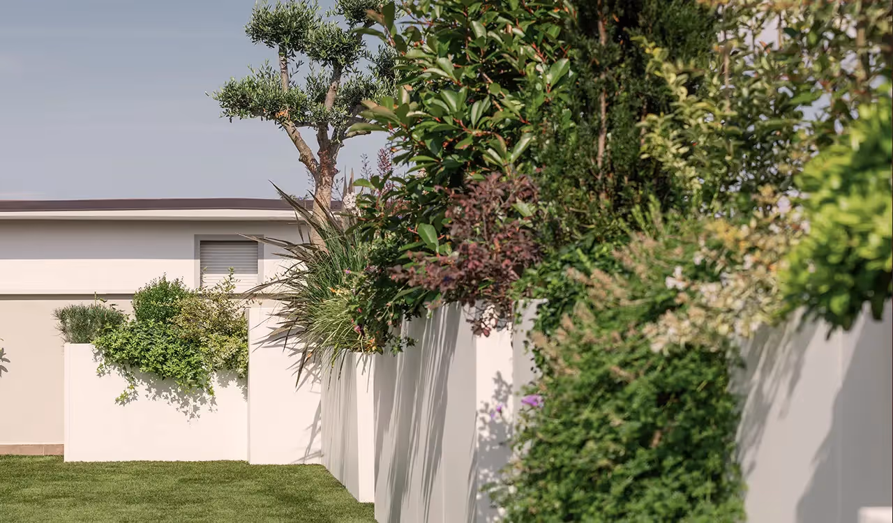 Vista laterale e ravvicinata di grandi fioriere modulari bianche con piante verdi e cespugli rigogliosi, posizionate lungo il bordo di un giardino con prato.