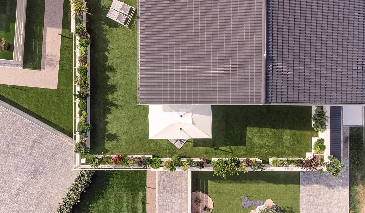Vista aerea zenitale di un tetto giardino a forma di L con prato sintetico, ombrellone bianco e una fila continua di fioriere perimetrali fiorite lungo la ringhiera.